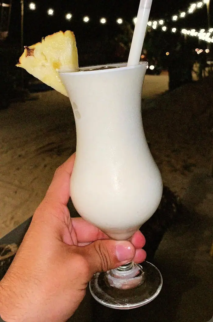 Piña colada servida na taça com fatia de abacaxi, segurada na mão à beira da praia