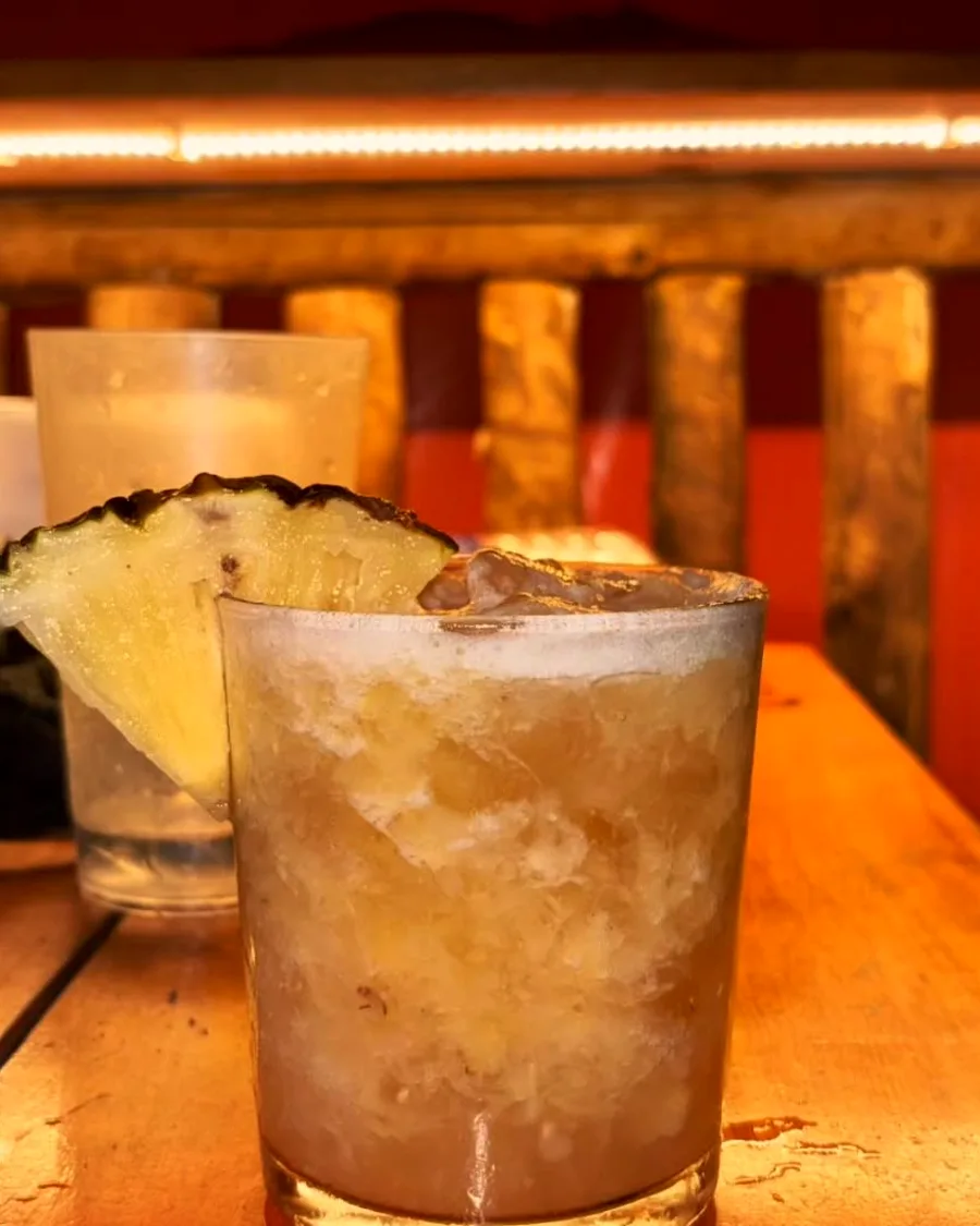 Dois drinks servidos na mesa de madeira, com a luz quente do salão atrás