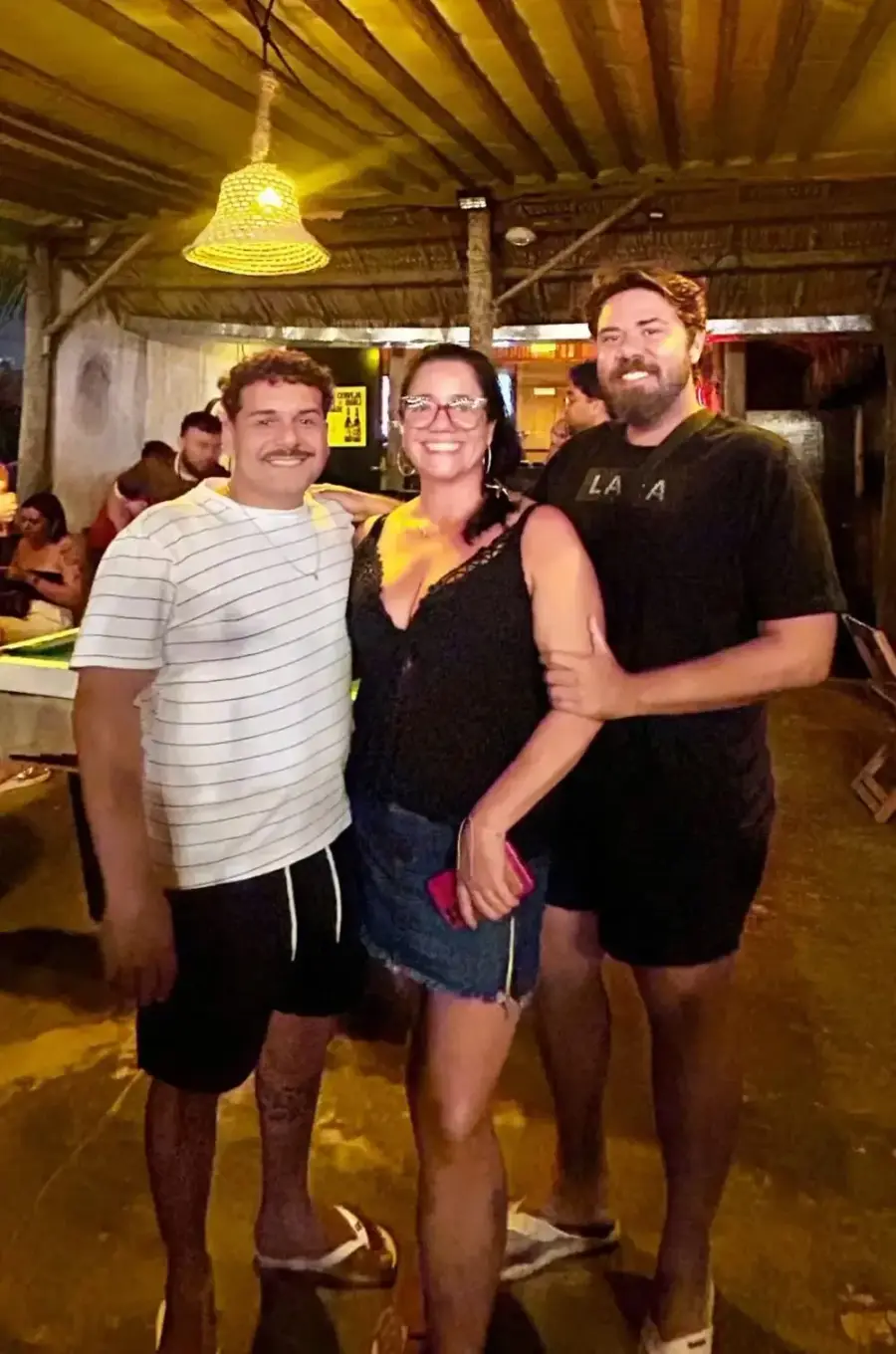 Três amigos abraçados no salão do Chalé, com o balcão iluminado ao fundo