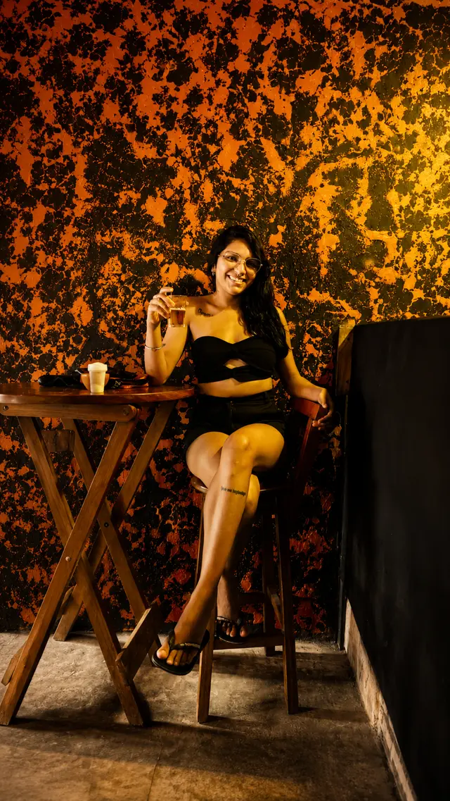 Cliente sentada na mesa alta do Chalé com um drink, diante da parede laranja texturizada