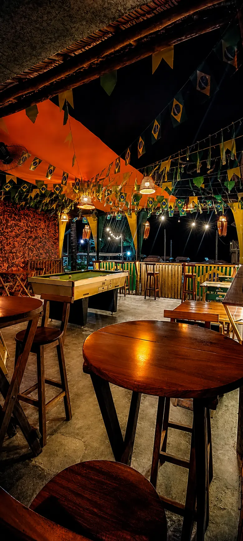 Mesas altas de madeira e a mesa de sinuca do Chalé, sob o toldo laranja e as bandeirinhas