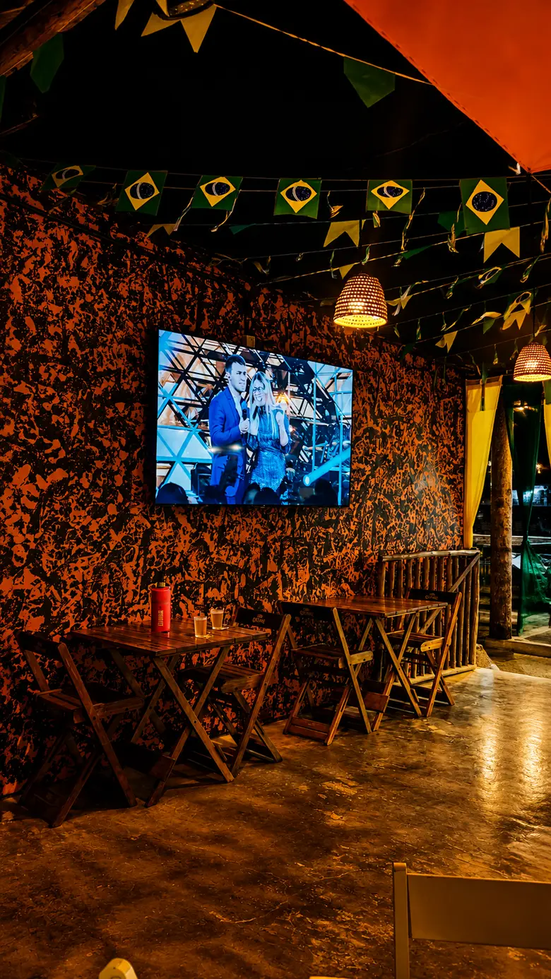 Canto do salão com TV na parede laranja, mesas de madeira e bandeirinhas no teto