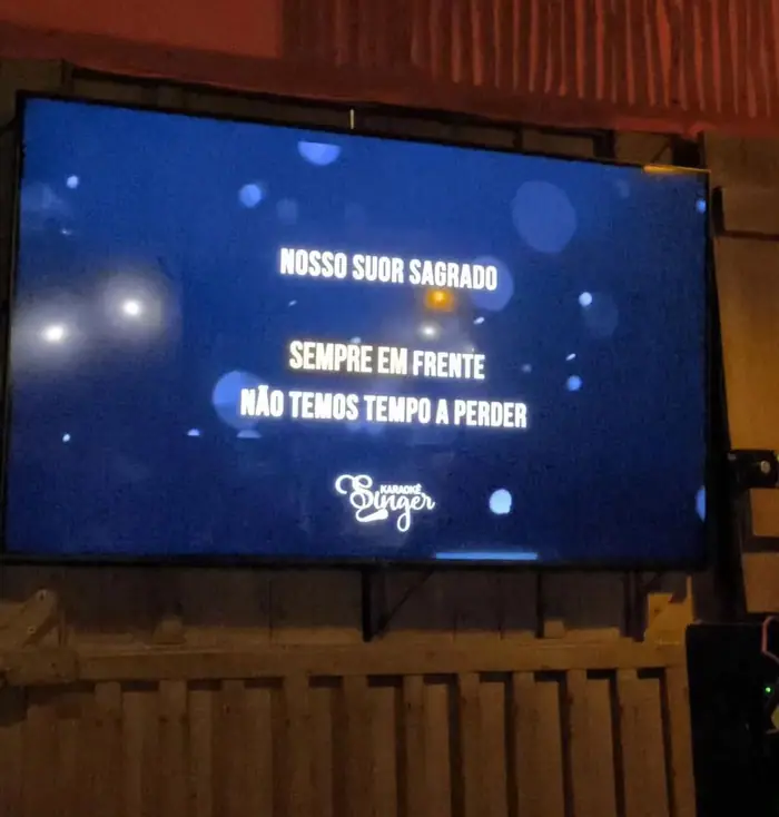 Telão do karaokê com a letra da música na tela, preso na parede de madeira