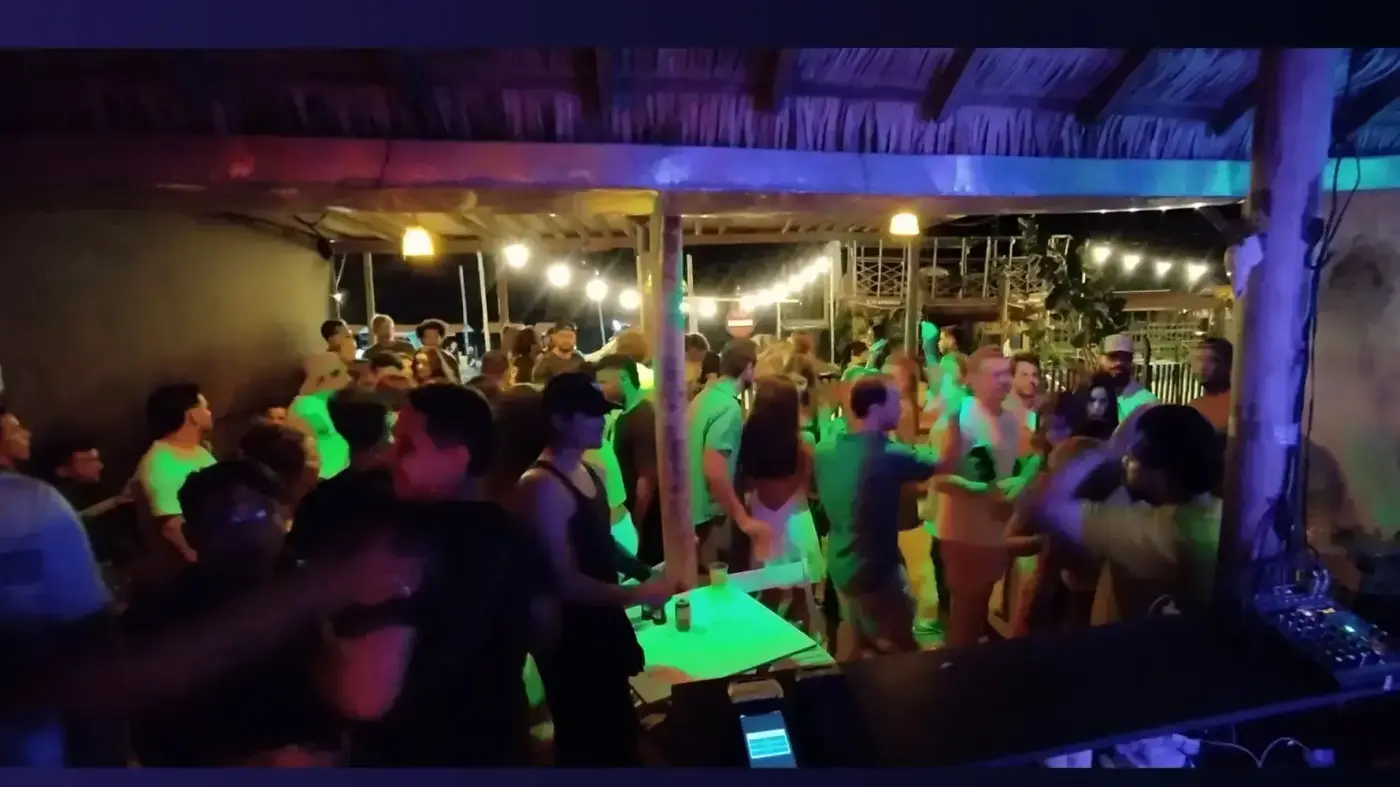 Salão lotado à noite sob luz verde, com as luzinhas do teto de palha acesas
